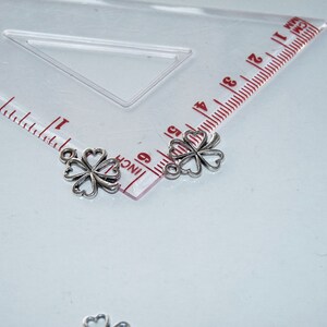 May include: Two silver four-leaf clover charms with a loop at the top. The charms are about 1 inch in size and are shown next to a ruler.