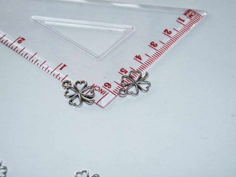 May include: Two silver four-leaf clover charms with a loop at the top. The charms are about 1 inch in size and are shown next to a ruler.
