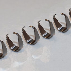 Puede incluir: Siete anillos de salto de metal plateados, también conocidos como anillos de salto o anillos de conexión, están dispuestos en una fila sobre una superficie blanca. Los anillos están abiertos y listos para ser utilizados para la fabricación de joyas.