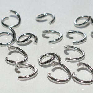 May include: A collection of small, shiny, silver-colored metal jump rings. Some rings are open, while others are closed. The rings are arranged on a white surface, and the lighting creates highlights on their curved surfaces.