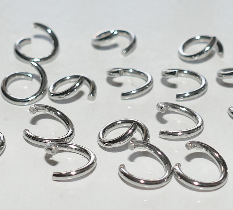May include: A collection of small, shiny, silver-colored metal jump rings. Some rings are open, while others are closed. The rings are arranged on a white surface, and the lighting creates highlights on their curved surfaces.