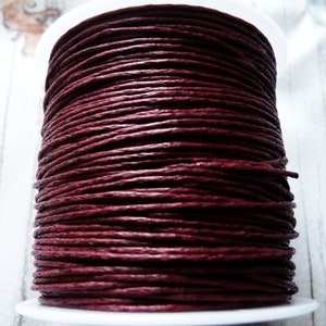 Puede incluir: Una bobina de cordón de algodón encerado de color rojo oscuro. El cordón está enrollado firmemente alrededor de una bobina de plástico blanco.