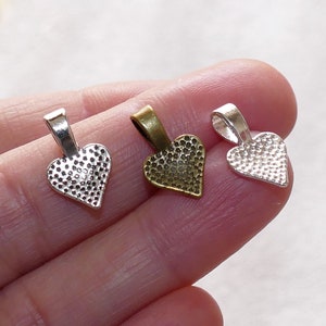 May include: Three heart-shaped metal pendant bails in silver, bronze, and gold tones. Each bail has a textured surface and a loop at the top for attaching a pendant.