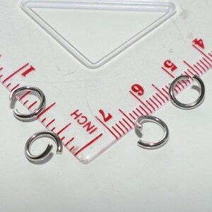 May include: Close-up of four small, silver-colored jump rings. The rings are open, with a small gap. A ruler with inch markings is in the background.