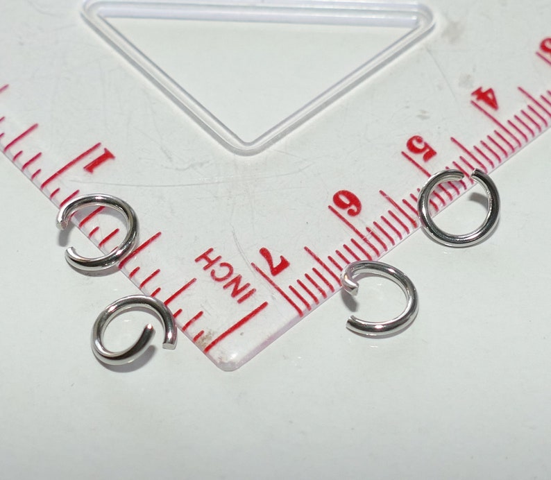 May include: Close-up of four small, silver-colored jump rings. The rings are open, with a small gap. A ruler with inch markings is in the background.