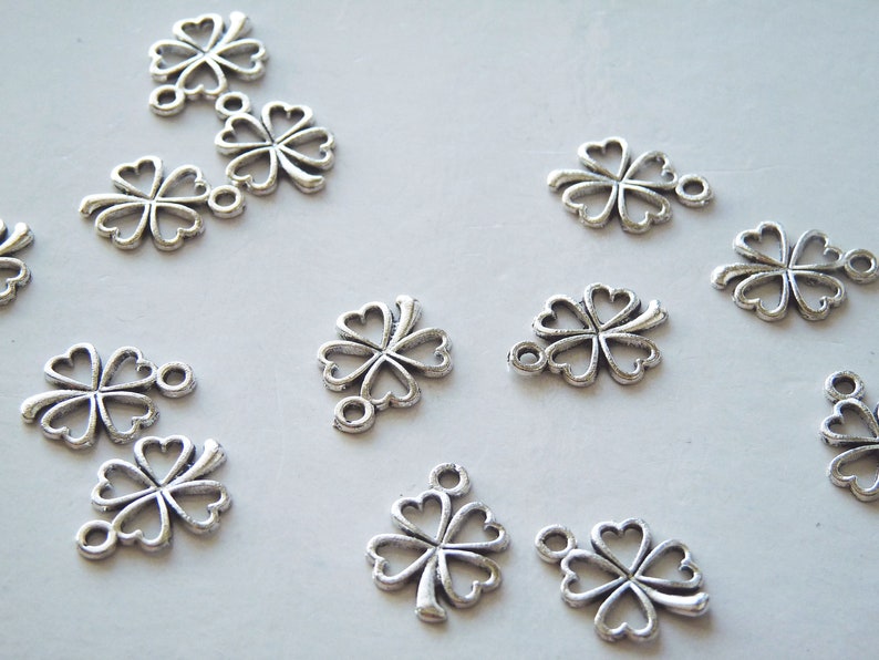 May include: A collection of silver-toned metal charms shaped like four-leaf clovers. Each charm has a loop at the top for attaching to jewelry or other crafts.