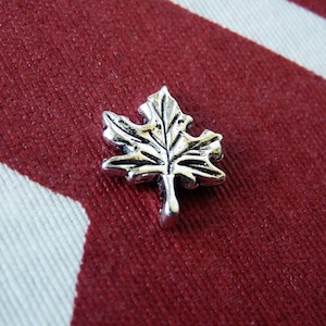 May include: A silver maple leaf pin on a red and white striped fabric background.