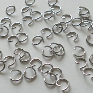 May include: A large quantity of silver jump rings, a type of jewelry finding, are scattered on a white surface. The rings are all the same size and are made of metal.