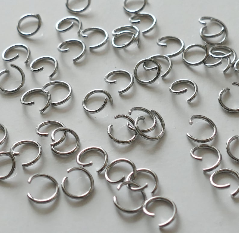 May include: A large quantity of silver jump rings, a type of jewelry finding, are scattered on a white surface. The rings are all the same size and are made of metal.