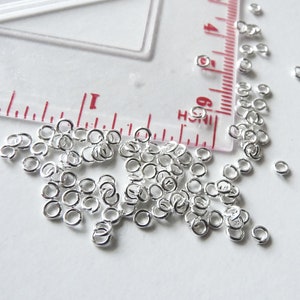 May include: A large quantity of silver jump rings, scattered on a white surface. The rings are small and round, with a smooth finish.
