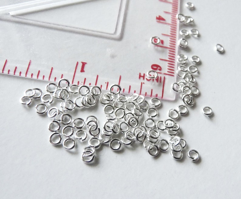 May include: A large quantity of silver jump rings, scattered on a white surface. The rings are small and round, with a smooth finish.