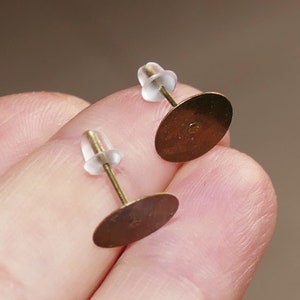 May include: Two earring posts with brass-colored oval pads and clear plastic backings. The posts are held between fingers, showing the detail of the earring components. The pads are slightly domed and have a small hole in the center.
