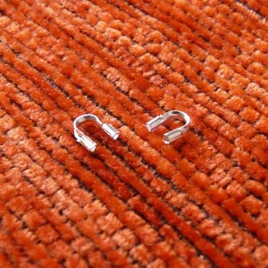 May include: Two small, silver-colored U-shaped metal connectors with rectangular ends are displayed against a textured, orange background. The connectors are positioned near each other, suggesting they are related.