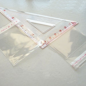 May include: Two clear plastic bags with red printed text. One bag has the text "Resealable" and the other has the text "This flap to open". A clear plastic ruler with red markings is on top of the bags.