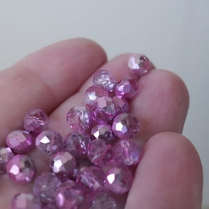 May include: A close-up of a hand holding a pile of iridescent pink glass beads. The beads are faceted and have a shimmery finish.