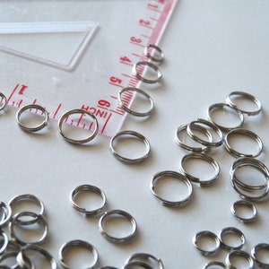 May include: A large quantity of silver jump rings of various sizes. The rings are scattered on a white surface.