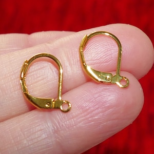 May include: Two gold-toned earring hooks, also known as leverback earring findings, are shown on a finger. The hooks are made of metal and have a smooth, polished finish.