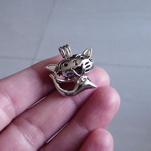 May include: Silver charm in the shape of a cat with a hollow belly. The cat has purple and red accents on its face and a small loop at the top for attaching to a necklace or bracelet.