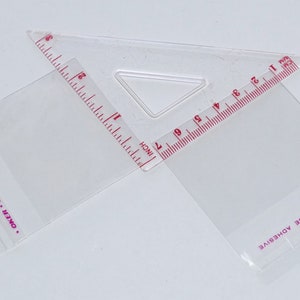 Puede incluir: Una regla de pl&aacute;stico transparente con forma de tri&aacute;ngulo y una regla a cada lado. La regla est&aacute; marcada en pulgadas y cent&iacute;metros. El texto en la regla dice "OVE LINER TO EXPOSE ADHESIVE" y "OKER BRAND SEALING".