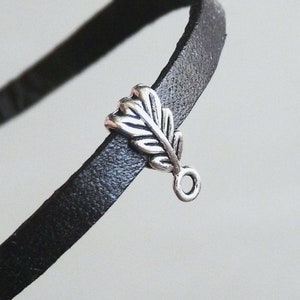 Peut inclure: Breloque en forme de feuille argentée avec une petite boucle en bas. Le charme est attaché à une sangle en cuir noir.