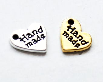 May include: Two heart-shaped metal charms, one silver-toned and one gold-toned, each featuring a small circular hole at the top and the black embossed text "Hand made" across the center of the heart surface.