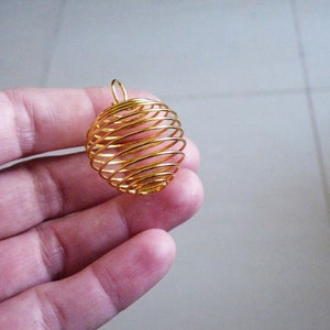 Può includere: Un ciondolo a forma di gabbia di filo dorato, un globo decorativo con un motivo a spirale, tenuto in una mano.