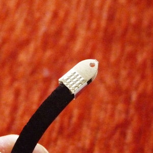 以下が含まれることがあります： 黒いコードのブレスレットに銀色の留め具が付いている。留め具の上部に小さな穴が開いている。