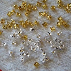 Könnte beinhalten: Eine Sammlung von goldenen und silbernen Metall-Quetschperlen für die Schmuckherstellung. Die Perlen sind klein und haben an einem Ende eine Schlaufe und am anderen Ende eine Quetschverbindung.