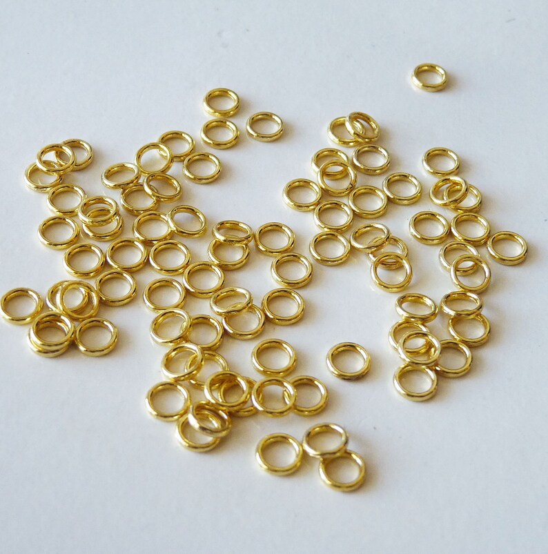 Puede incluir: Una gran cantidad de anillos de salto dorados, esparcidos sobre una superficie blanca. Los anillos son peque&ntilde;os y circulares, con un acabado liso y brillante.