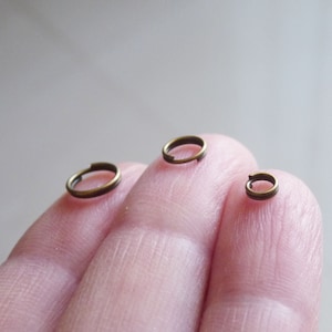 May include: Three gold jump rings of different sizes, shown on a finger.