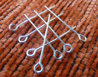 May include: A group of eight silver-toned head pins with loops on one end. The pins are arranged in a criss-cross pattern on a brown textured surface.