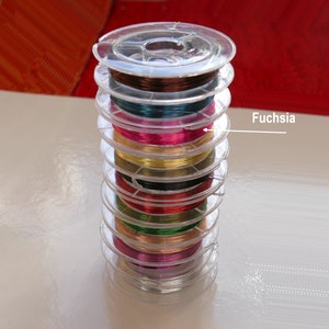 Può includere: Una pila di bobine di filo colorato, tra cui fucsia, oro e verde. Le bobine sono disposte a forma di torre, con la bobina fucsia in cima.