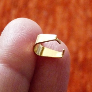 Puede incluir: Un pequeño hallazgo de joyería de color dorado, probablemente una fianza o cierre, se sostiene entre los dedos. La pieza de metal tiene un diseño curvo y abierto con un extremo en forma de gancho. El fondo es de color naranja sólido.