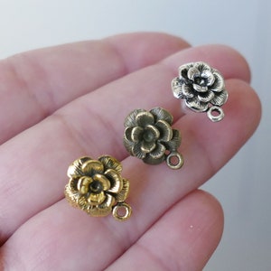 May include: Three flower-shaped metal charms with a loop for attaching to jewelry or other crafts. The charms are in gold, silver, and bronze.