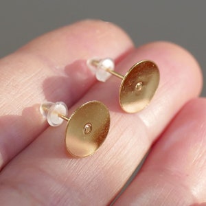 May include: Two gold-toned earring backs with a round, flat surface and a small hole in the center. The backs are attached to clear plastic posts.
