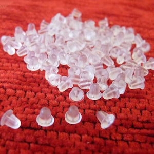 Puede incluir: Un mont&oacute;n grande de respaldos de pendientes de pl&aacute;stico transparente sobre una superficie roja texturizada.