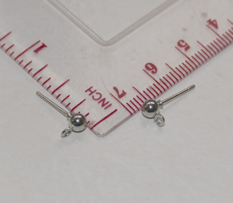 May include: Two silver earring findings with a small ball at the end and a loop for attaching jewelry. The findings are measured against a ruler.