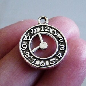 Op de afbeelding: Zilveren bedel in de vorm van een wijzerplaat met Romeinse cijfers en een zilveren wijzer. De bedel heeft een gat aan de bovenkant om aan een ketting of armband te bevestigen.