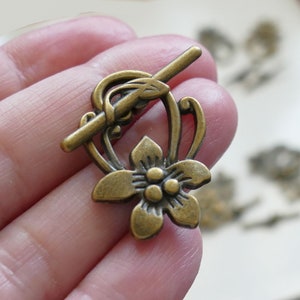 May include: Antique gold-tone metal toggle clasp with a floral design. The clasp features a delicate flower with three round accents in the center and a leaf detail on the top.