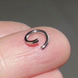 May include: A small, silver-colored, open ring, likely jewelry, is shown. The ring is curved, with a gap at one end. The ring is resting on a person's fingertip, with a neutral background.