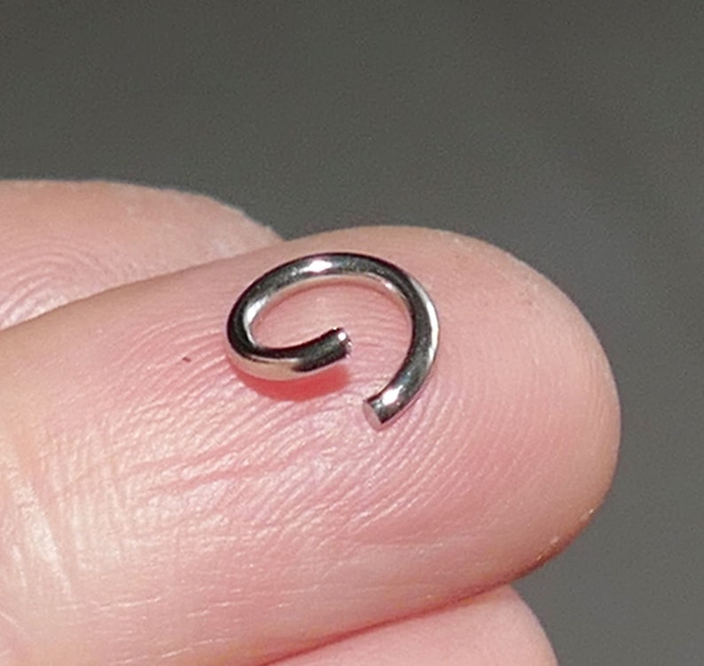 May include: A small, silver-colored, open ring, likely jewelry, is shown. The ring is curved, with a gap at one end. The ring is resting on a person's fingertip, with a neutral background.