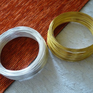 Può includere: Due bobine di filo, una argentata e una dorata, sono appoggiate su una superficie arancione testurizzata. Le bobine sono entrambe rotonde e hanno un diametro di circa 7,5 cm.