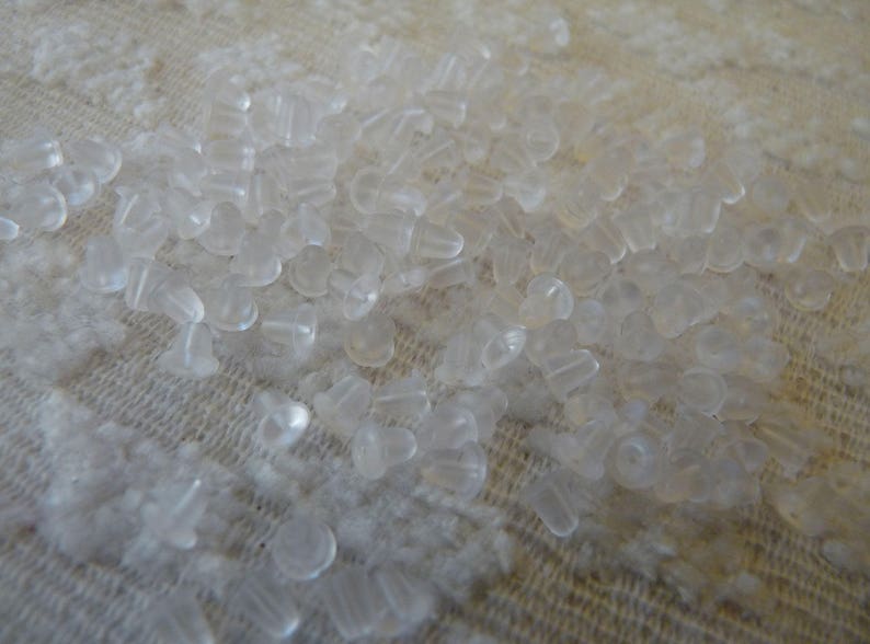 Puede incluir: Una gran cantidad de respaldos de pendientes de pl&aacute;stico transparente esparcidos sobre una superficie de tela blanca.