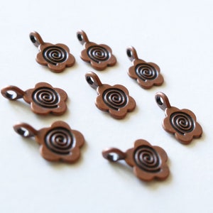 Op de afbeelding: Acht koperkleurige metalen bedels met een swirl-ontwerp in het midden. Elke bedel heeft een lus aan de bovenkant om te bevestigen aan sieraden of andere knutselwerkjes.