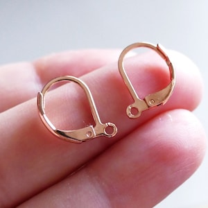 May include: Two rose gold earring hooks, also known as leverback earring findings, are shown on a hand. The hooks are made of metal and have a smooth finish.