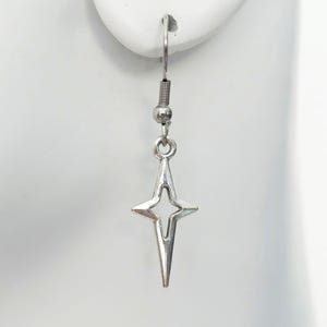 Op de afbeelding: Een zilverkleurige hanger-oorbel met een vierpuntige sterbedel met een hol centrum. De bedel hangt aan een eenvoudige zilverkleurige vishaak-oorhaak, wat zorgt voor een strakke en metalen uitstraling die verticaal vanaf de oorlel hangt.