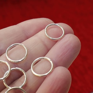 May include: A close-up of a hand holding several silver jump rings. The rings are small and circular, and they are perfect for jewelry making or other craft projects.