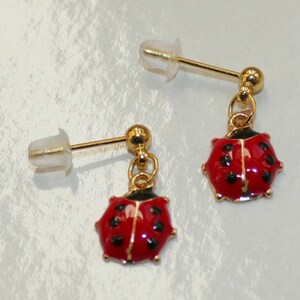 Könnte beinhalten: Ein Paar goldene Marienkäfer-Ohrringe mit roter und schwarzer Emaille. Die Ohrringe haben einen Stiftverschluss.