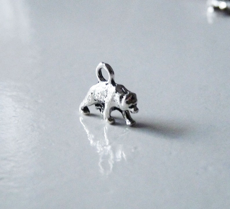 May include: Silver charm in the shape of a bear standing on all fours. The bear is facing to the right and has a loop at the top for attaching to a necklace or bracelet.