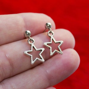 Può includere: Orecchini pendenti a forma di stella color argento. Ogni orecchino presenta un piccolo perno rotondo e un design a stella aperta pendente. Gli orecchini sono mostrati su uno sfondo rosso.